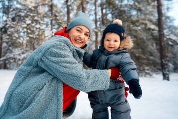 Fototapeta premium Young mother and her baby playing in the snowy forest. Young mother carrying her baby in the forest on a very snowy day.