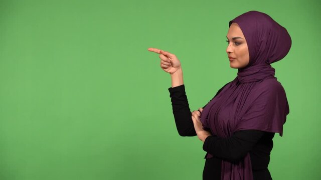 A young beautiful Muslim woman points at a bullet-point text and looks at the camera - green screen background