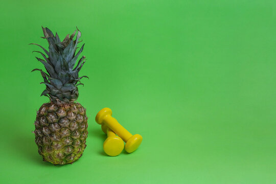 Sports Background, Dumbbells And Pineapple.
Sports Equipment And Healthy Food On A Green Background. View From Above. Motivation.