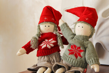 Textile toy figures of a boy and a girl in New Year's themed clothes and hats, in red-gray-white colors, sit on a shelf. Christmas and New Year decoration.