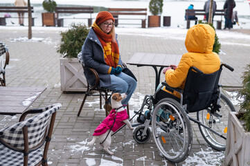 Two girlfriends in a cafe on a street terrace in winter. Woman in a wheelchair.