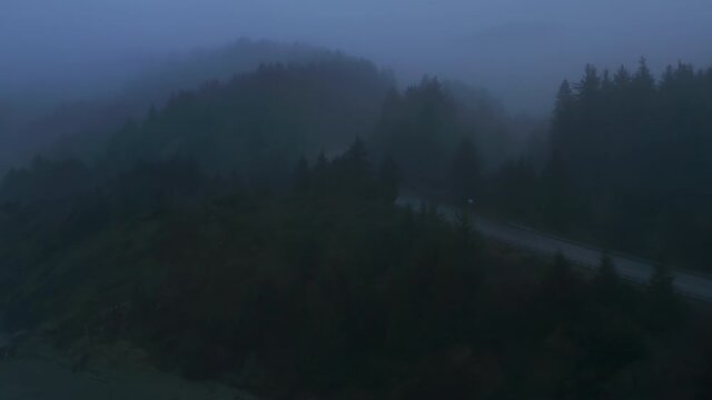 Aerial view of the grayish skyline above the picturesque highway with cars driving along. Dense, lush forest surrounds a coastal road with thick fog above. High quality 4k footage