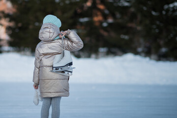 Fototapeta na wymiar A little girl in a blue hat and winter jacket looks at the ice of the lake and spruce, and holds white skates over her shoulder on a frosty winter day. Back view. Active weekend getaway in winter
