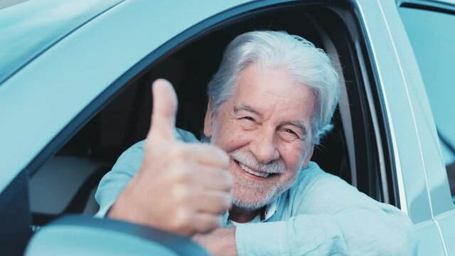 Happy owner looking at the camera with happy face and thumbs up. Handsome bearded mature man sitting relaxed in his newly bought car looking out the window smiling joyfully. One old senior driving and