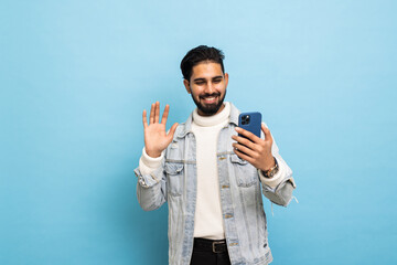 Handsome young Asian man with a smartphone is making a video call, isolated on blue background