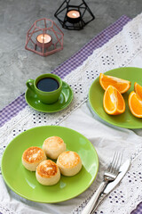 Breakfast cheesecakes, oranges and cup of coffee in light green dishes with candles on grey table