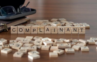 complainant concept represented by wooden letter tiles on a wooden table with glasses and a book