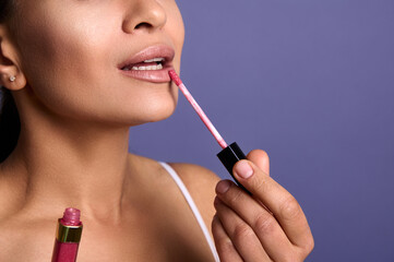 Close-up of an attractive young woman with perfect clean skin applying lip gloss or hygienic balm on plump lips isolated on purple background with copy space for advertisement. Make-up concept