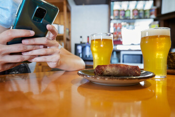 Anonymous person with a mask taking a photo with a smartphone of the appetizer consisting of butifarra sausage and a couple of beers