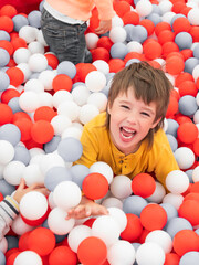 Fototapeta na wymiar Laughing boy plays in ball pit. White and red plastic balls in dry paddling pool. Leisure activity in playroom at kindergarten. Leisure activity. Active recreation.