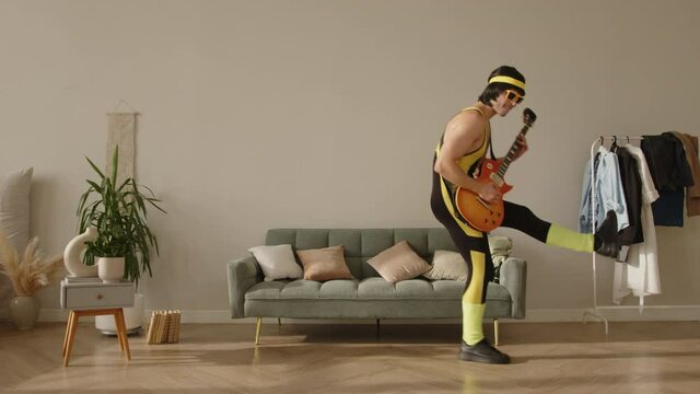 Young funny hipster guy at home dressed in a vintage tight yellow tracksuit with sunglasses and a guitar in his hands dancing, playing and posing like a rock star. Home entertainment concept