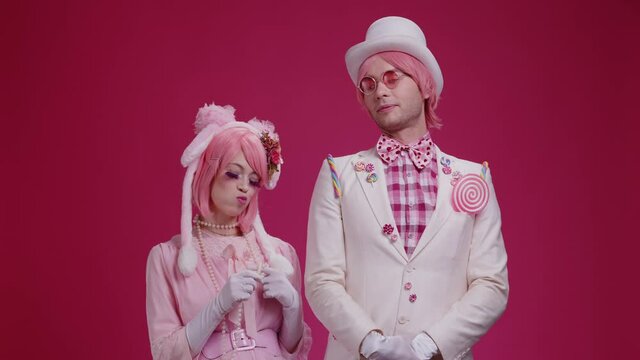 Couple of colorful actors mimes in pink colored costumes stand on pink studio background, a guy flirts with a girl, gives a lollipop, a girl kisses a guy on the cheek. Cute love scene
