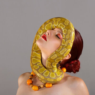 A beautiful young woman poses for the camera. 
Her necklace is made of kumquats fruit and a 
yellow python snake curls itself around her face.
