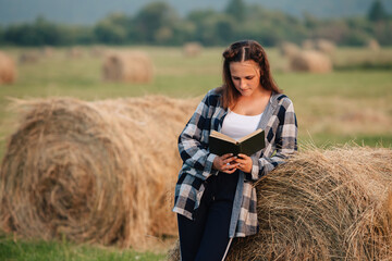 A young adult girl is reading a book in a field with her elbows on a roll of hay.