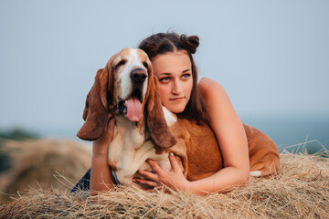 A young adult girl walks with a Basset Hound dog in nature. The owner hugs the pet.