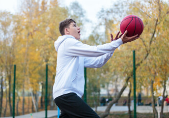 Fototapeta premium Cute teenager in white hoodie playing basketball. Young boy with red ball learning dribble and shooting on the city court. Hobby for kids, active lifestyle