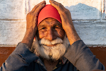 portrait of an old indian man, old aged man with wrinkles on his face holding his face with his hands and sitting in the sunlight.