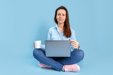 A young adult girl is sitting with a laptop on her lap and holding a white mug in her hands. Blue background with copyspace