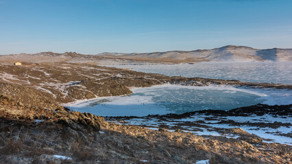 A small frozen lake in the shape of a heart and a large one next to it. There are tire tracks on the ice. Snow and dry grass on cold ground. A lonely house on a hill. Blue sky. Siberia