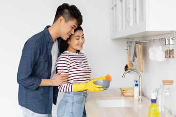 Asian guy hugging his cheerful girlfriend washing dishes at kitchen