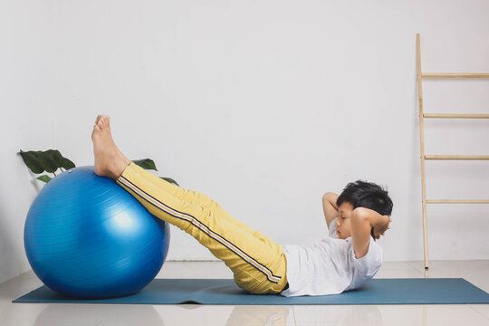 Active Asian Kid Doing Sit Ups With Feet On Gym Ball At Home