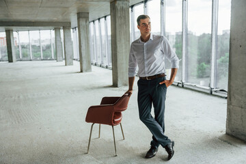 Near the chair. Young man in formal wear is working indoors on the construction