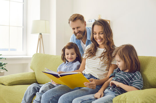 Happy mother, father and children reading together. Mum, dad and two kids sitting on comfortable sofa at home, looking through family photo album or book of funny interesting stories and fun facts