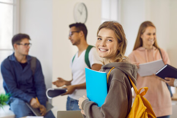Happy beautiful female university student with book and backpack. Portrait of pretty young girl with candid cheerful face expression smiling at camera while standing in classroom with her classmates - obrazy, fototapety, plakaty