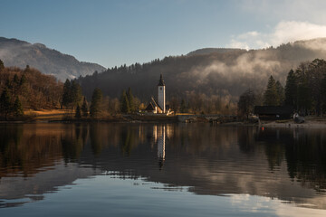 Atmospheric serene view of the white Catholic church reflecting in the water. Sunny morning. Bohinj lake. Slovenia.