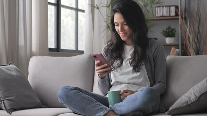 cheerful young arab woman sitting cross legged on the couch using smart phone mobile on internet,latin girl sits on the sofa holding smartphone searching online - Powered by Adobe