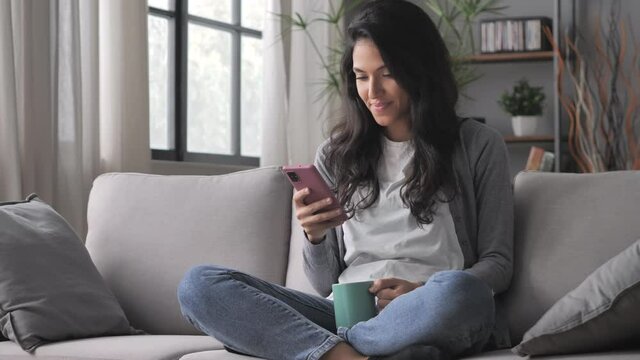 cheerful young arab woman sitting cross legged on the couch using smart phone mobile on internet,latin girl sits on the sofa holding smartphone searching online