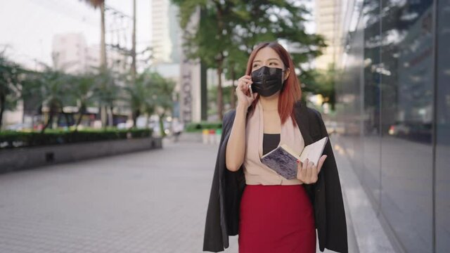 Woman office worker wear medical mask using mobile phone talking to clients, dealing with problem through the phone, a secretary carrying paper work documents, smart business attire, modern city urban
