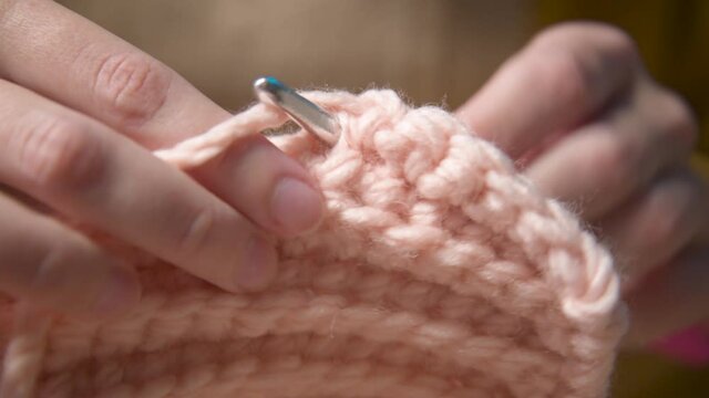Close-up. Hands of an attractive young caucasian woman in casual clothes sitting on a sofa indoors knits a woolen product by a crochet