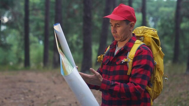 man tourist search route map. hiker with a backpack stands in the woods holding a paper map. adventure travel navigation concept. tourist in a red cap in a travel forest lifestyle with a backpack