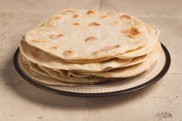 Indian bread (roti) on brown and beige plate are on beige stone table. Close-up.