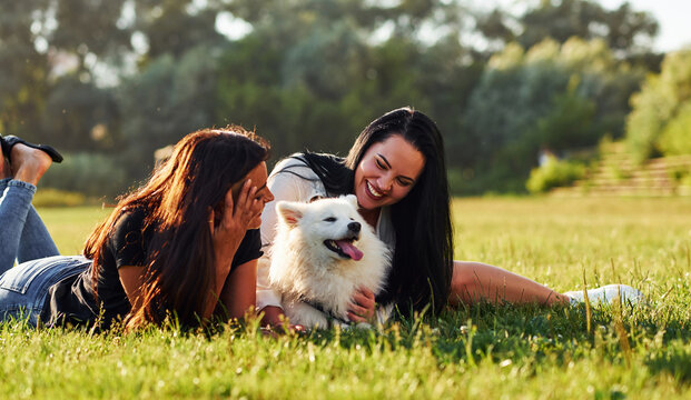 Two women having a rest while laying down on the ground of field with their cute dog
