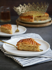 Pieces of traditional French galette des rois or king cake on a white plate with crown and glass of cider.A traditional cake usually served on Epiphany, made with puff pastry and creamy almond filling