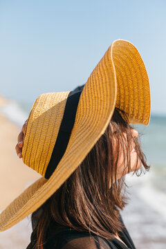 Portrait of carefree happy woman with windy hair in hat relaxing on sunny beach at sea. Summer vacation. Stylish calm young female in straw hat enjoying day on tropical island. Tranquil moment