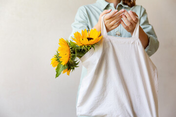 Eco-natural cotton reusable bag with sunflowers, hanging on the arm of a girl in a light green jumpsuit, on a light background. The concept of an ecological lifestyle and clothing. copy space