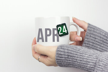 April 24th. Day 24 of month, Calendar date. Closeup of female hands in grey sweater holding cup of tea with month and calendar date on teabag label. Spring month, day of the year concept.