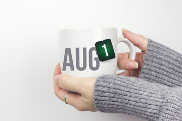 August 1st . Day 1 of month, Calendar date. Closeup of female hands in grey sweater holding cup of tea with month and calendar date on teabag label. Summer month, day of the year concept.