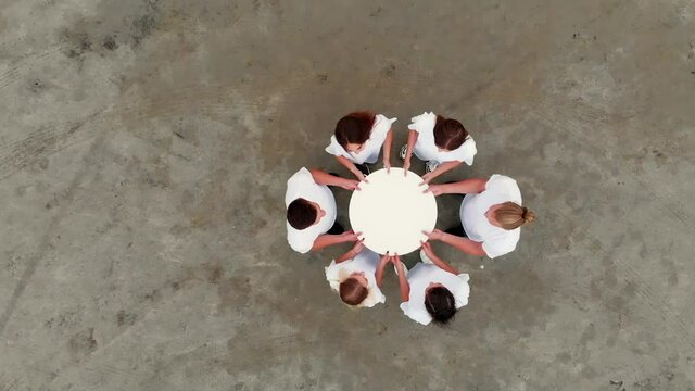 Group of male and female friends wearing jeans and white t-shirts holds edges of light paper circle together standing on ground aerial view