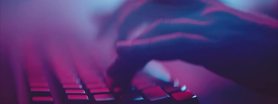 Low angle shot of fingers pressing keys on keyboard. Dark interior lit by dim colour light. Shallow depth of field. 