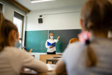 School teacher during corona virus pandemic wearing face mask and teaching pupils in classroom.
