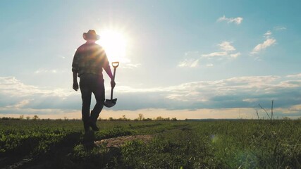 Farm agriculture concept. Farmer with a shovel in rubber boots walks across field at sunset. Silhouette man gardener walking along the road along the field. Tired man after hard work on the ground.