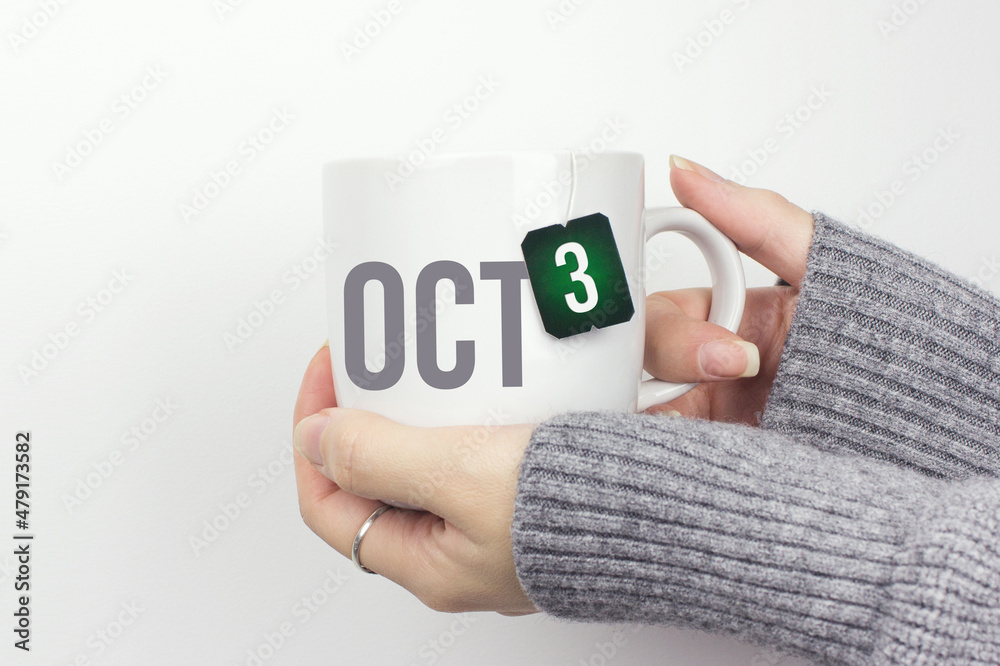 Wall mural October 3rd. Day 3 of month, Calendar date. Closeup of female hands in grey sweater holding cup of tea with month and calendar date on teabag label. Autumn month, day of the year concept.