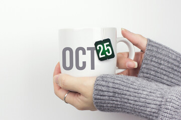 October 25th. Day 25 of month, Calendar date. Closeup of female hands in grey sweater holding cup of tea with month and calendar date on teabag label. Autumn month, day of the year concept.