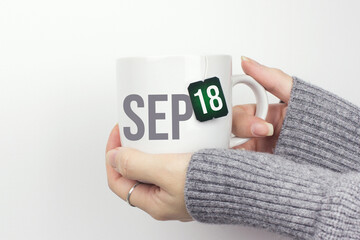 September 18th. Day 18 of month, Calendar date. Closeup of female hands in grey sweater holding cup of tea with month and calendar date on teabag label. Autumn month, day of the year concept.