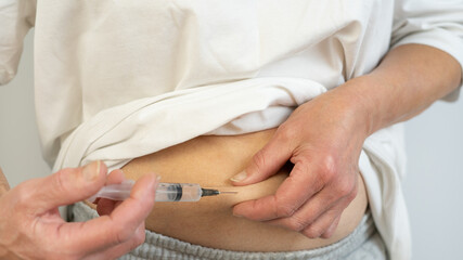 A woman in light clothes, giving herself an injection in the stomach, on a white background. He holds a syringe with medicine in his hand. Vaccination. An injection. Virus protection. Close-up.