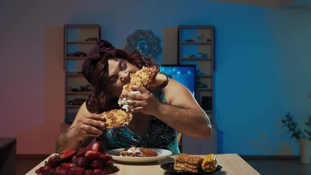 A man dressed in a woman's dress, a wig with long hair and makeup on his face eats a huge sandwich sitting at a table with sweets and fruits.
